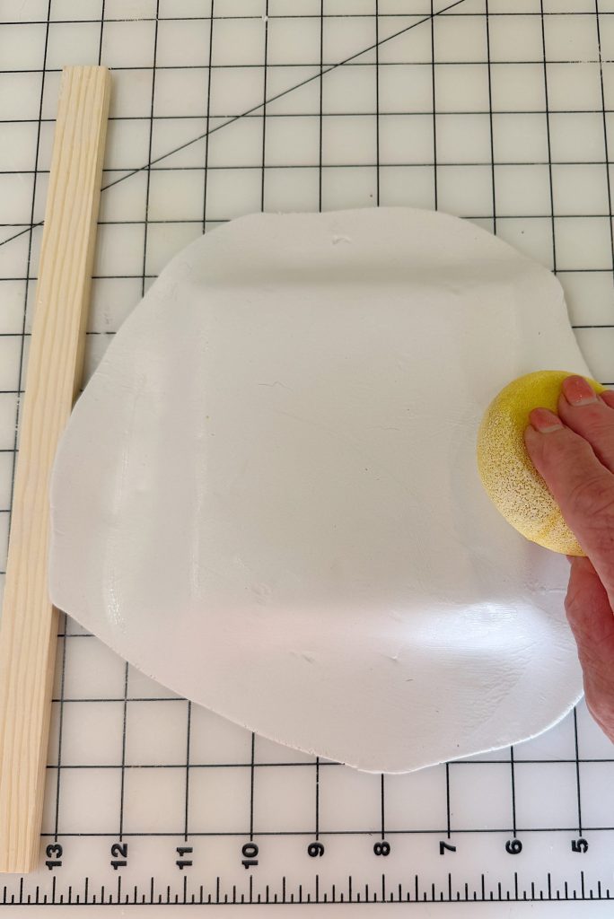 A hand holding a yellow sponge smooths a rolled-out piece of white clay on a gridded surface, with a wooden stick placed on the side.