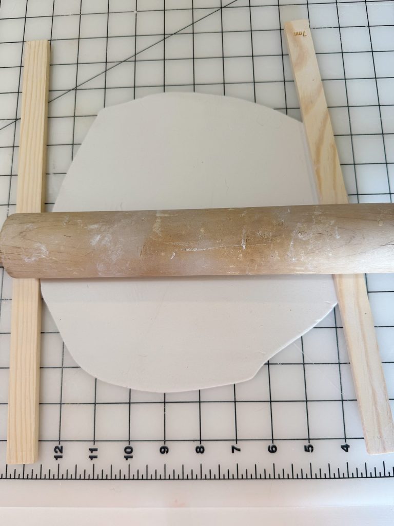 A rolling pin flattens white clay between two wooden guides on a gridded cutting mat.