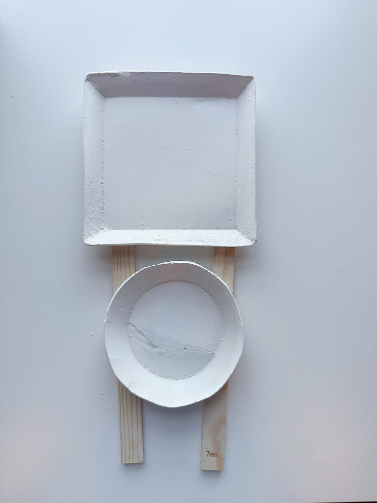 A square plate and a round bowl are placed on two wooden sticks against a white background, arranged to resemble a geometric face.