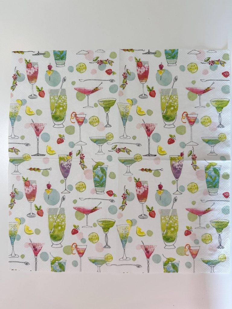 A patterned napkin featuring various colorful illustrated cocktails, garnished with fruit, and scattered with lemon and lime slices on a white background.
