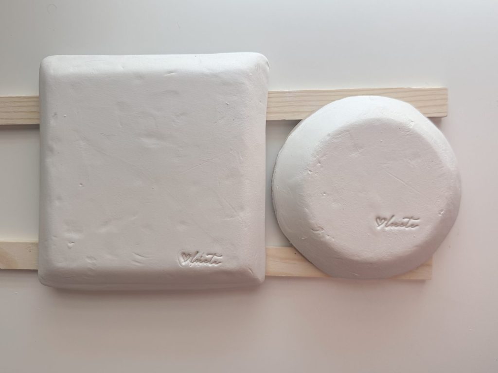 A square and a round slab of white clay, each imprinted with a small heart symbol and the word "date," rest on two parallel wooden slats on a white surface.