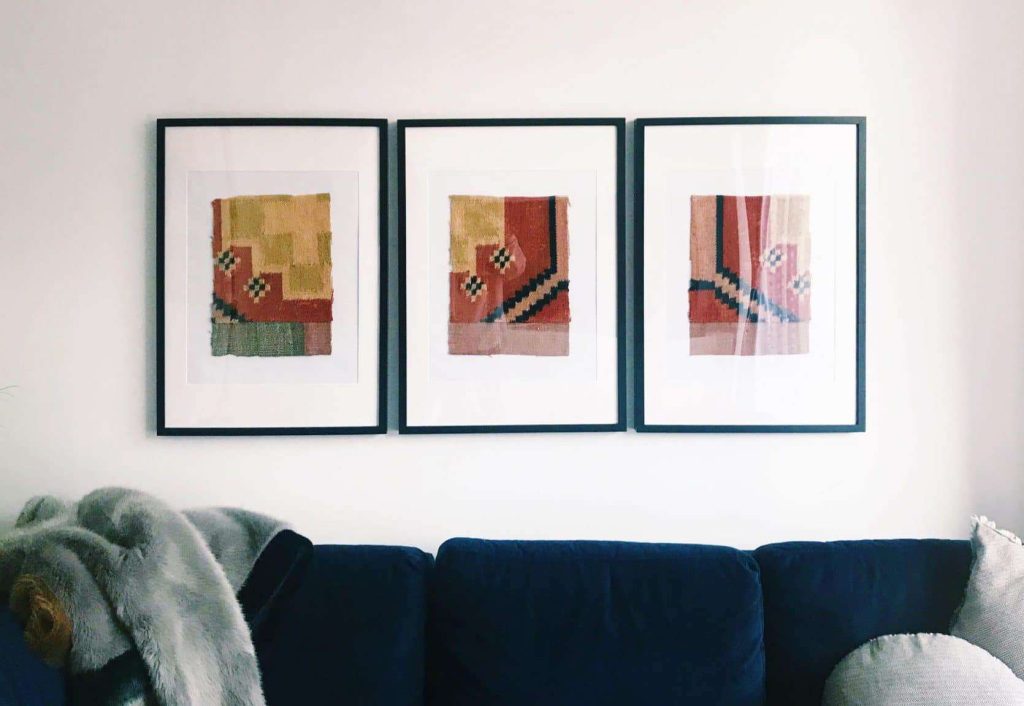 Three framed abstract artworks with geometric patterns hang in a row above a dark blue sofa; a light gray throw is draped over the sofa’s arm.