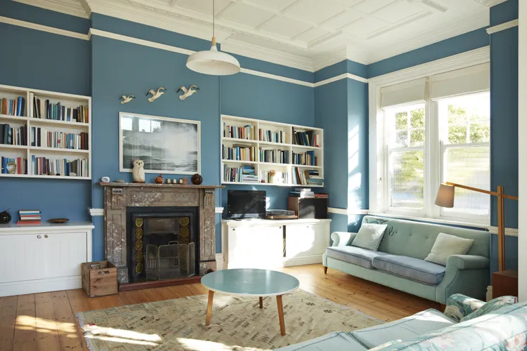 A living room with blue walls, built-in bookshelves, a fireplace, a round coffee table, sofa, armchair, and large windows letting in natural light.