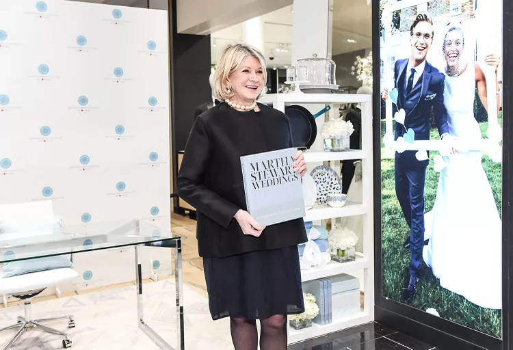 A woman in a black dress stands indoors holding a book titled "Martha Stewart Weddings," with wedding-themed decor and a large wedding photo display in the background.