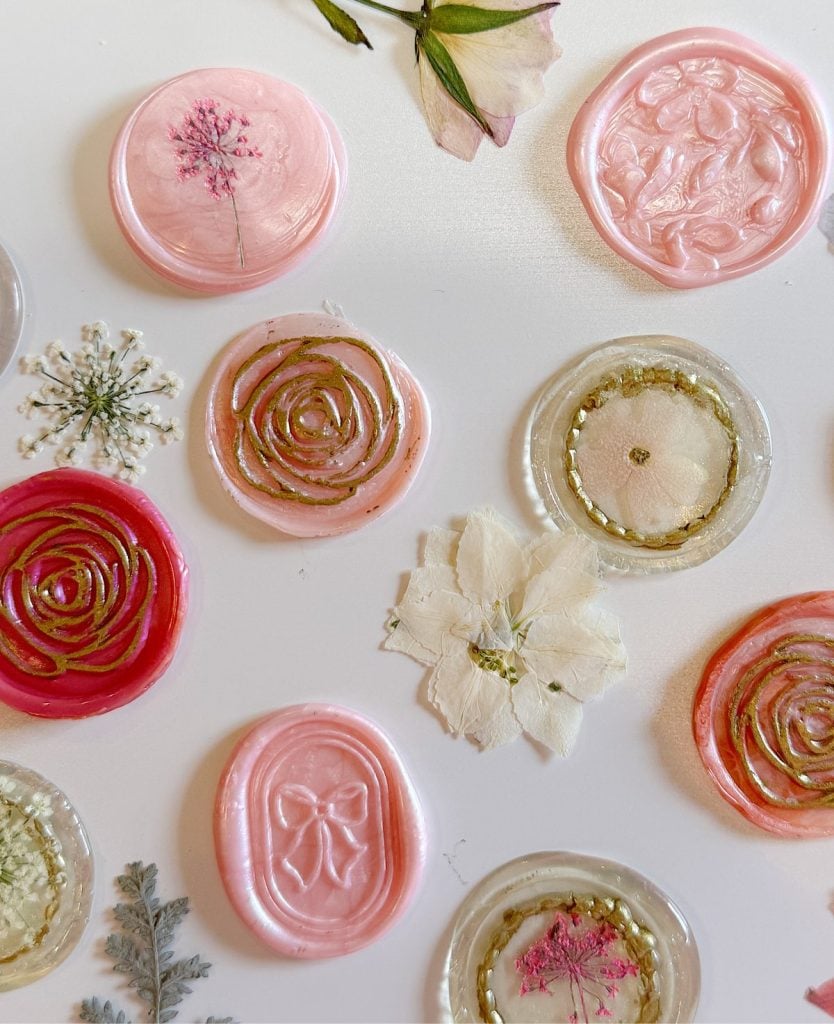 Assorted pink and gold wax seals, some with dried flowers embedded, are arranged on a white surface alongside pressed flowers.