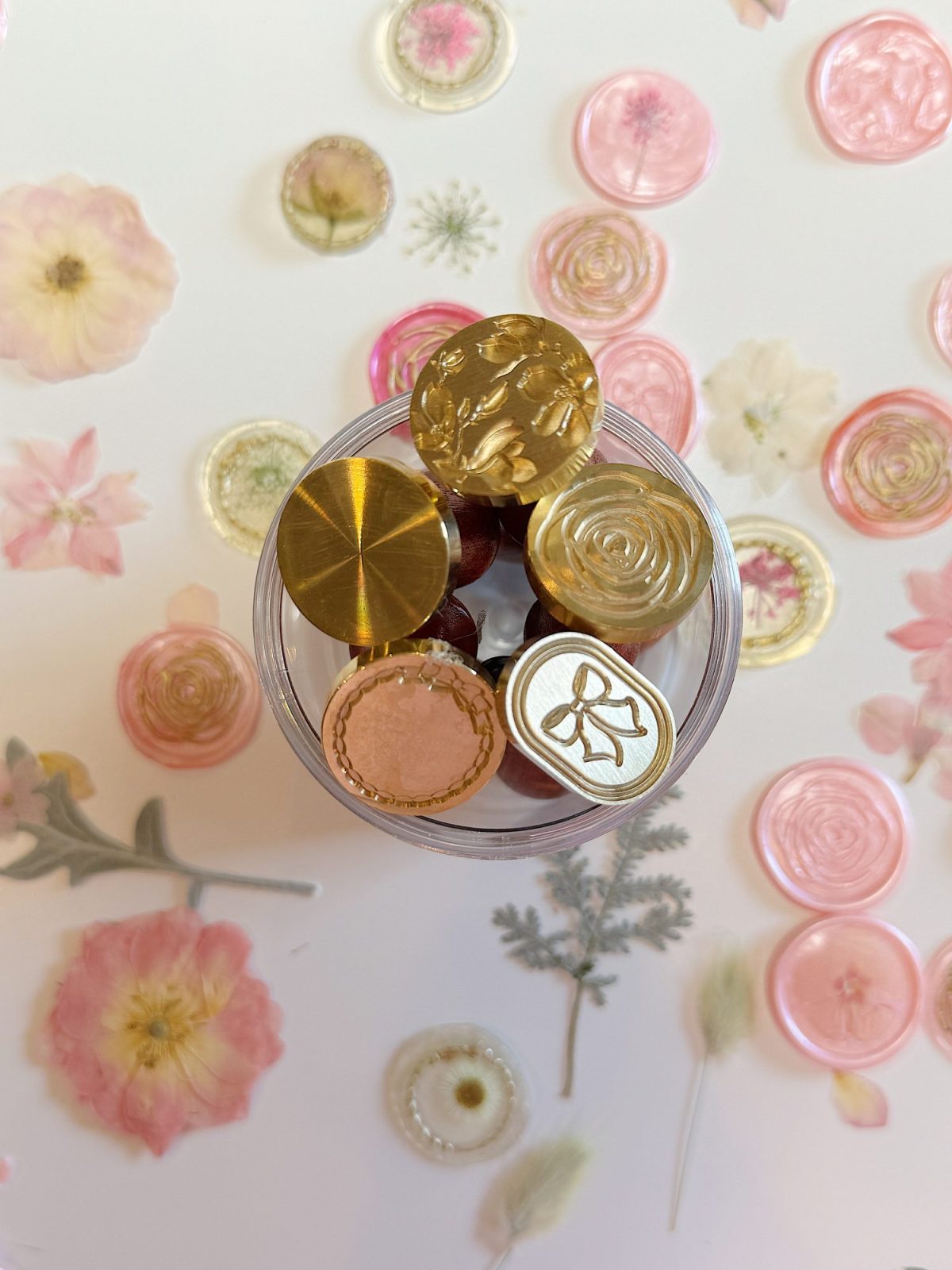 A clear container holds several brass wax seal stamps, surrounded by pastel pressed flowers and wax seals on a white surface.