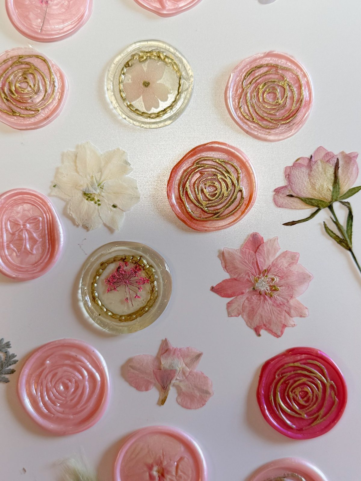 Assorted pink and gold wax seals alongside pressed and dried flowers arranged on a white surface.