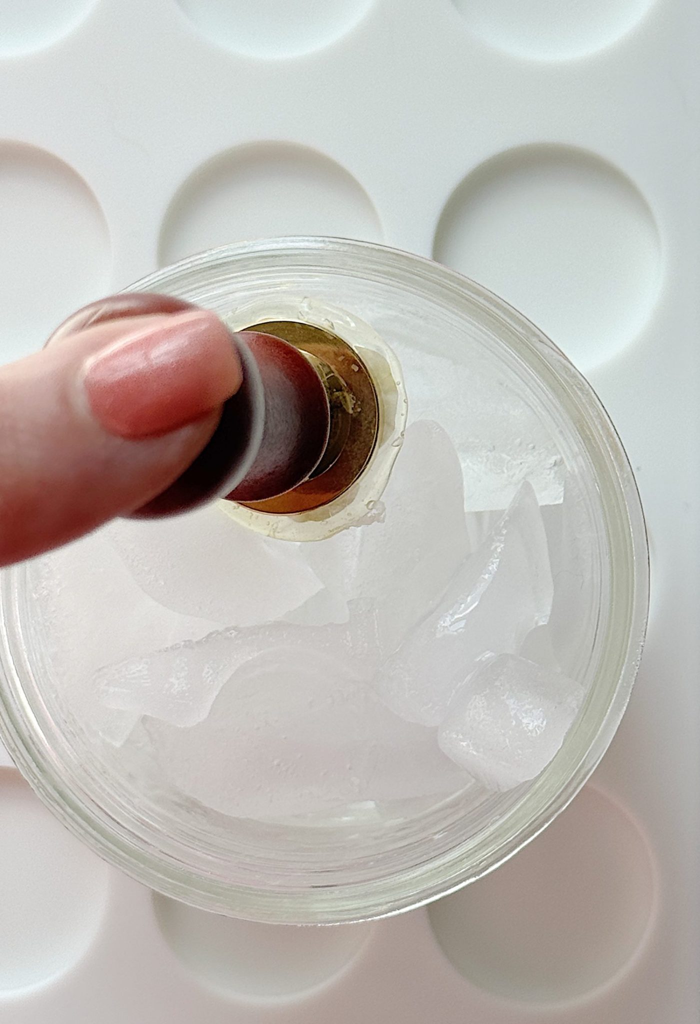 A hand dips a red lipstick tube into a glass filled with ice cubes, seen from above on a white surface with circular indents.