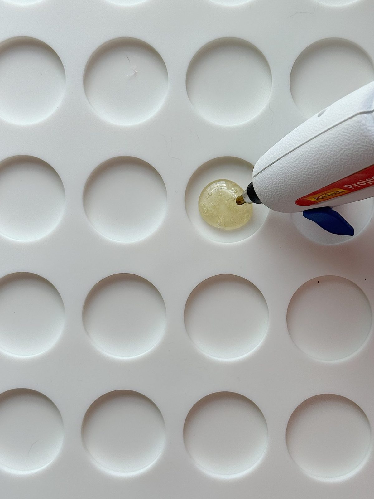 A hot glue gun applies a drop of hot glue onto one circular indentation of a white, dimpled surface.