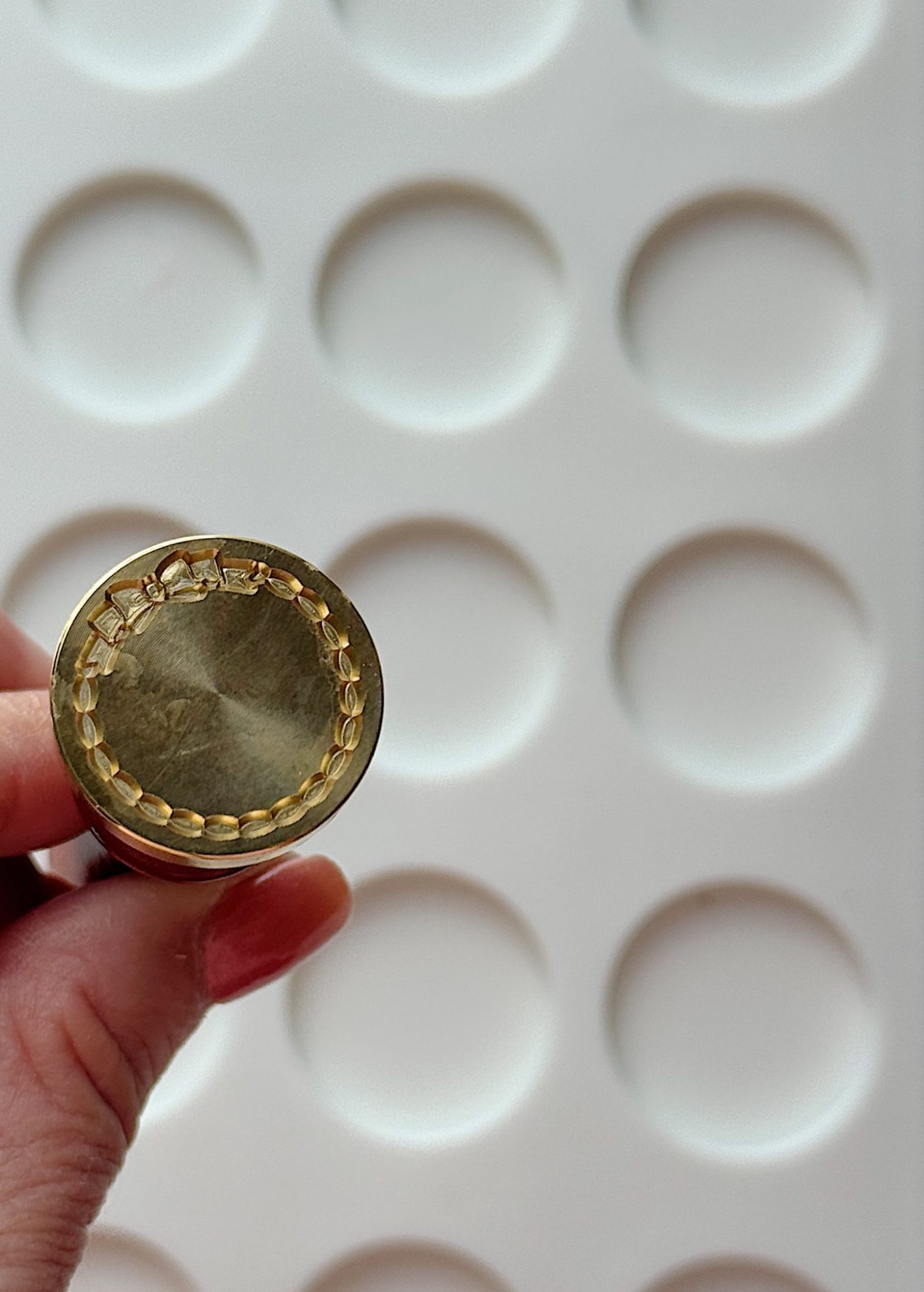 A hand holds a brass stamp with a circular pattern, in front of a white tray with round indentations.