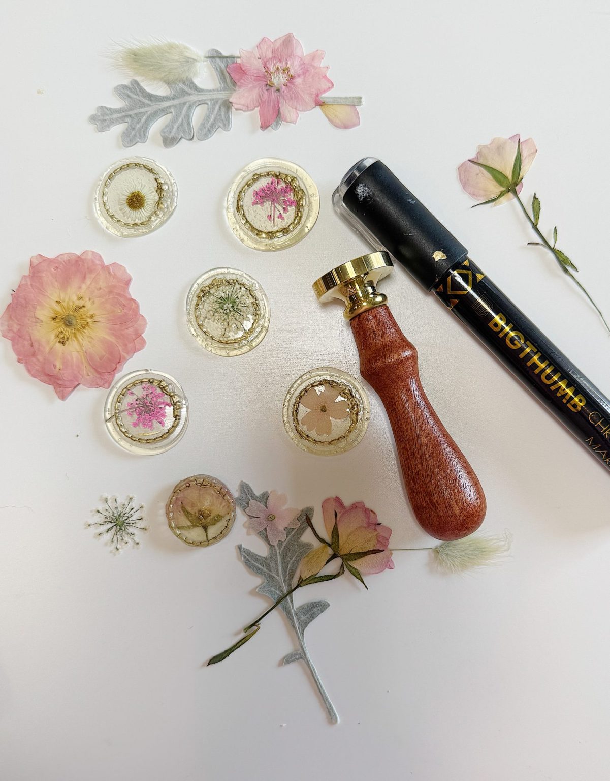 Dried flowers and leaves, wax seals with pressed flowers inside, a gold wax seal stamp, and a black sealing wax stick arranged on a white surface.