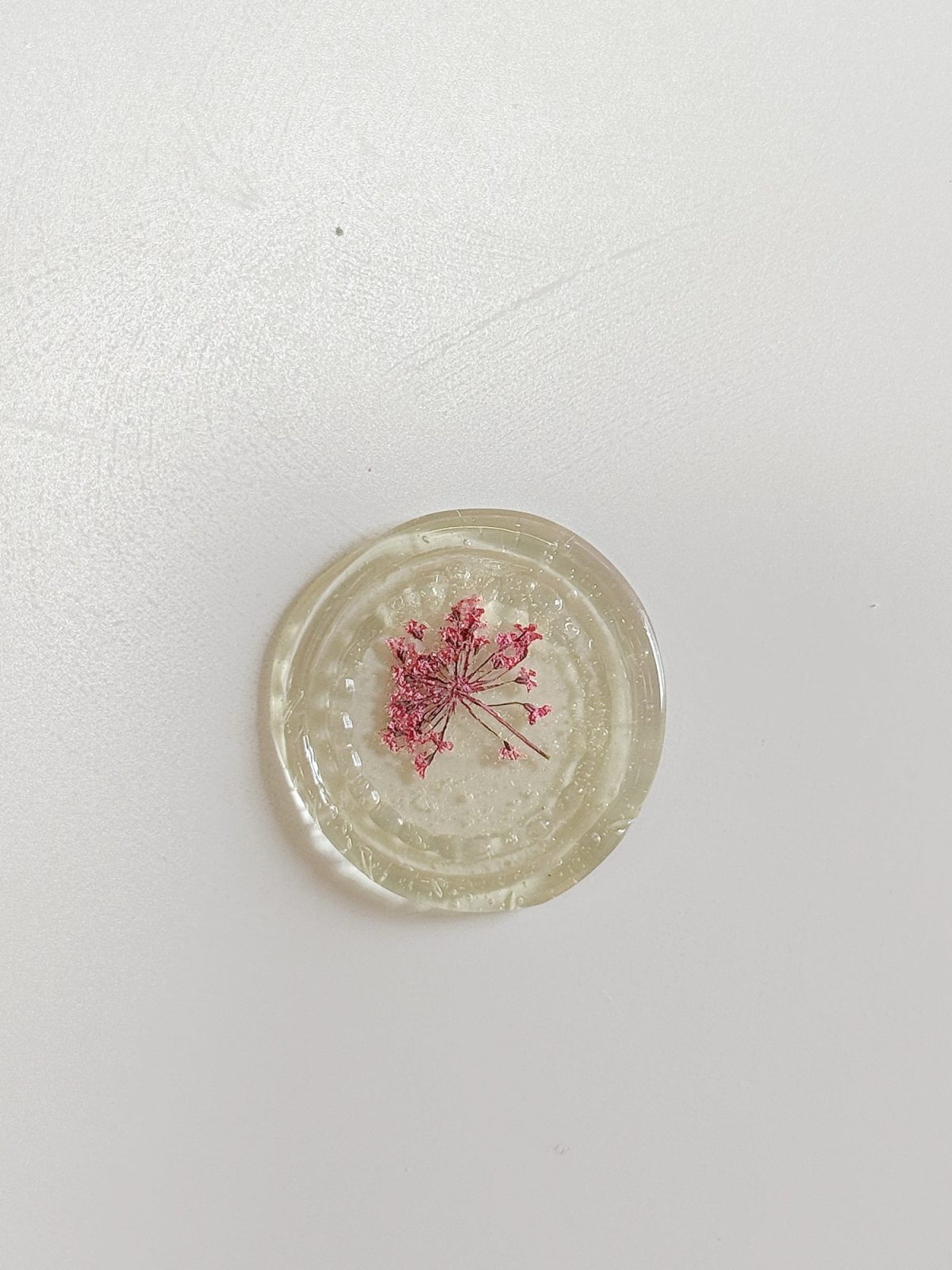 A round clear resin piece with a small pink dried flower embedded in the center, displayed on a plain white background.
