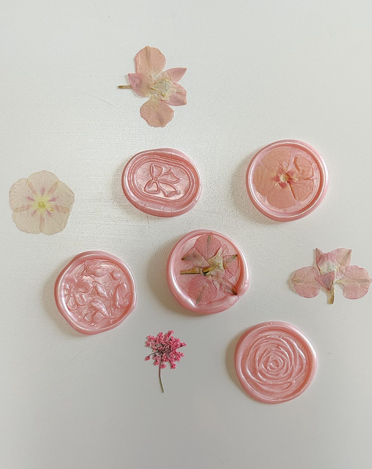 Five pink wax seals and several pressed pink flowers are arranged on a white surface. Each wax seal features a different floral design.