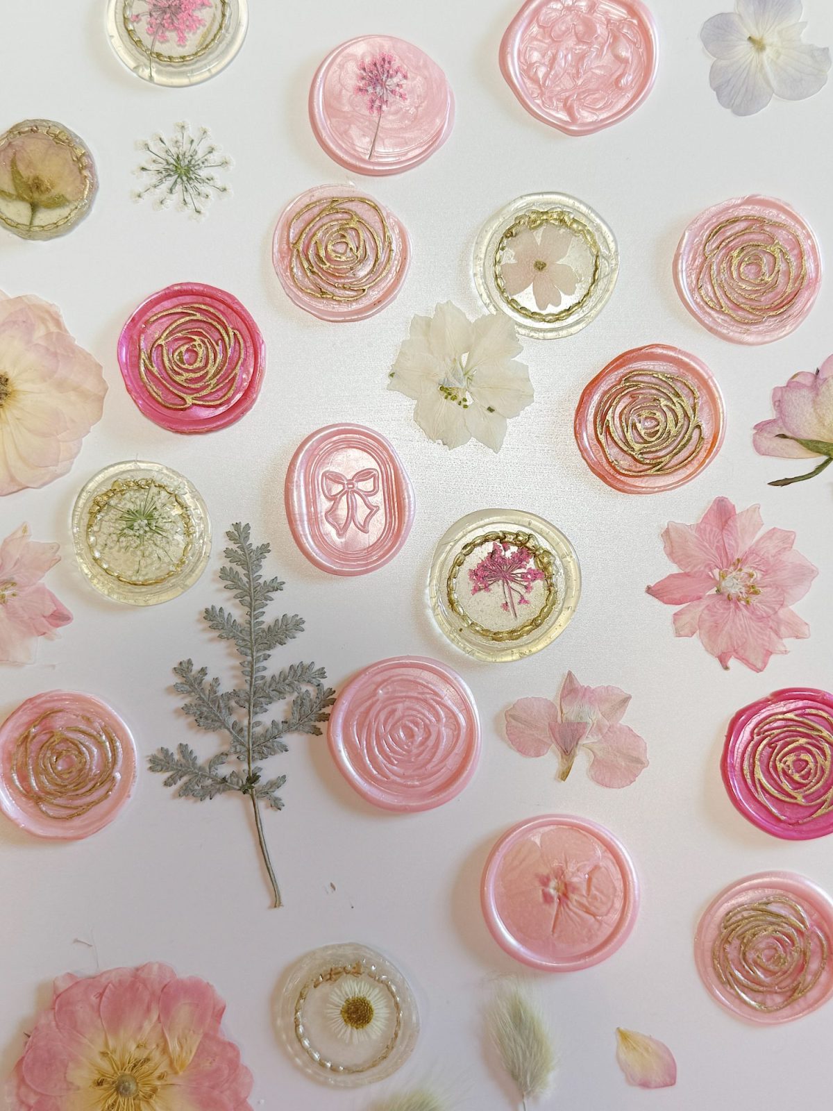 Pink and gold wax seals and assorted pressed flowers and leaves are arranged on a white background.