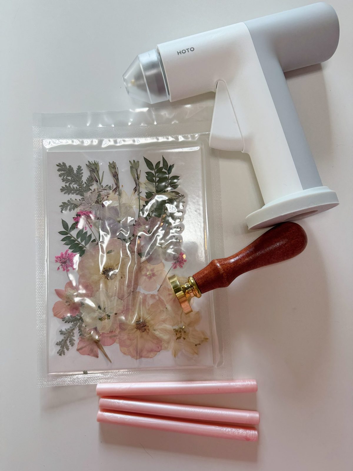 A heat sealer, a wax seal stamp with a wooden handle, three pink wax sticks, and a bag containing pressed flowers arranged on a white surface.
