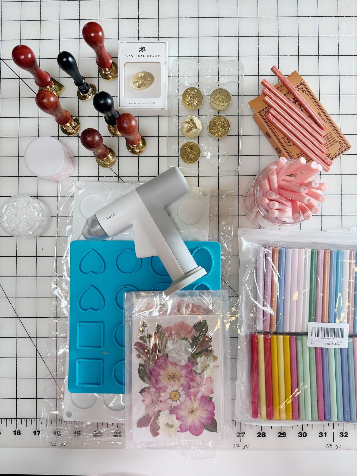 Crafting supplies arranged on a grid surface, including wax stamps, wax beads, a heat gun, molds, dried flowers, colored markers, and pink wax sticks.