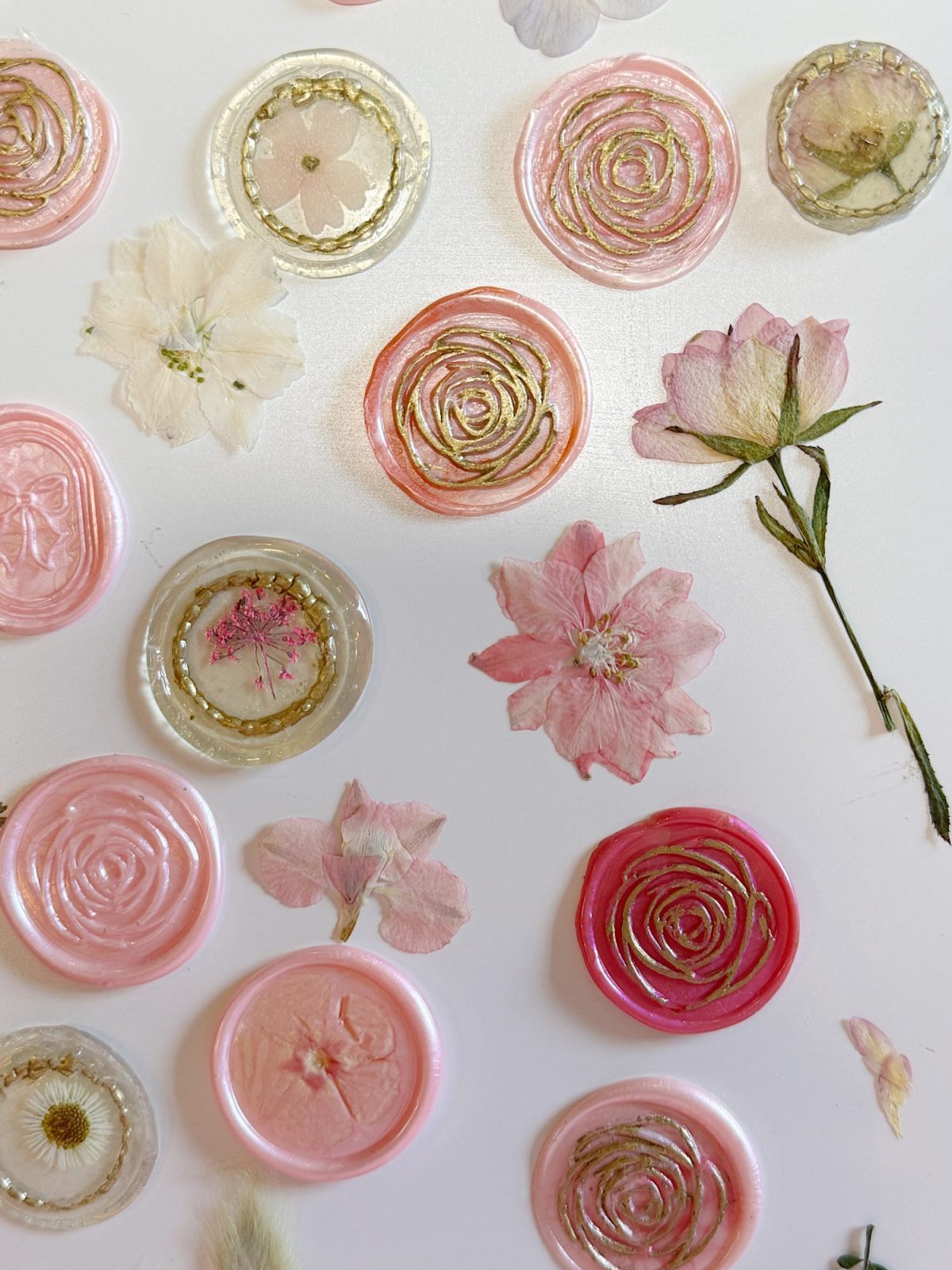 Pink and clear wax seals with gold accents are arranged on a white surface alongside various pressed pink and white flowers.