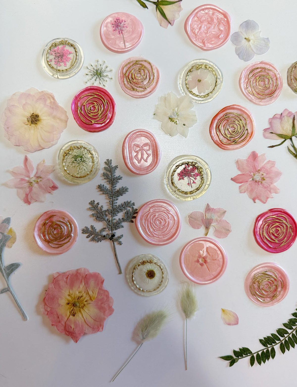 Assorted pink and white wax seals with floral designs are arranged on a white surface with dried flowers and leaves scattered among them.