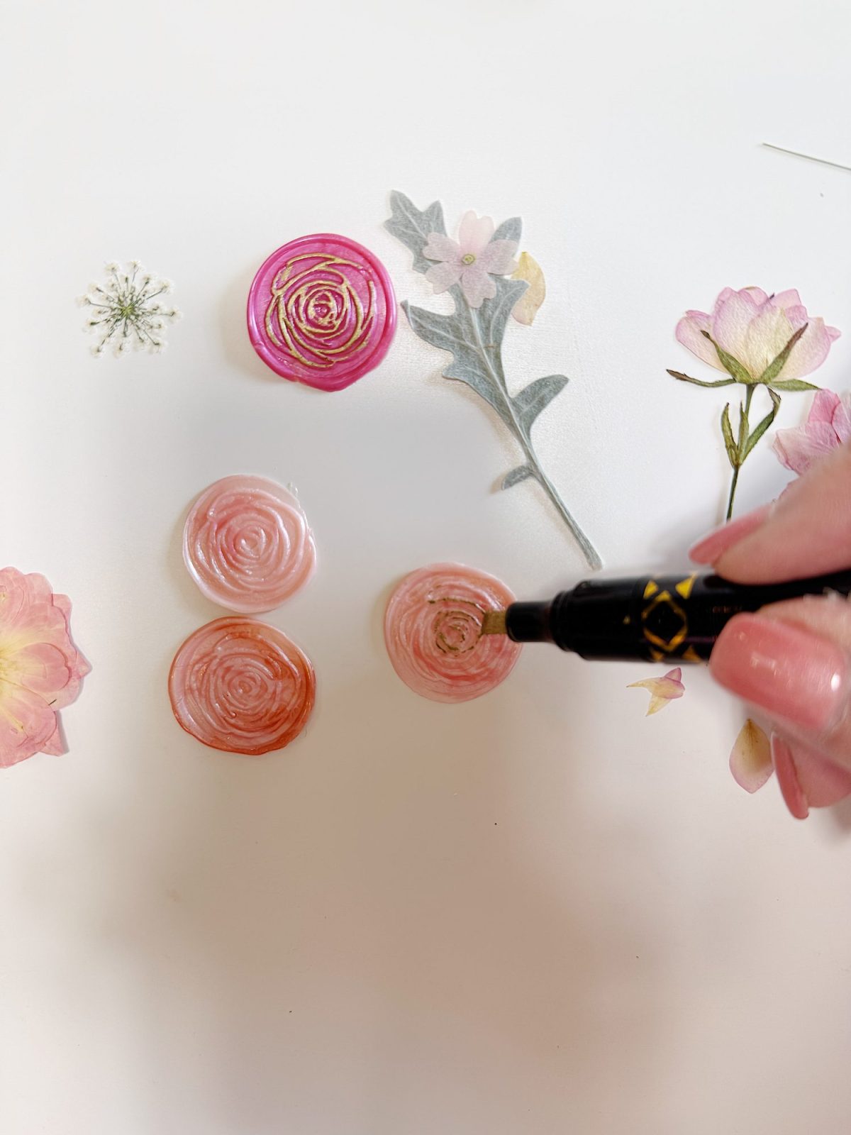 A hand uses a black marker to add details to pink circular flower designs surrounded by various paper flower cutouts on a white surface.