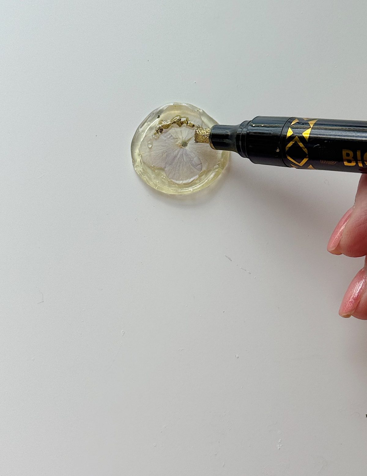 Hand holding a black and gold pen, writing on a clear resin piece with a pressed flower inside, placed on a white surface.