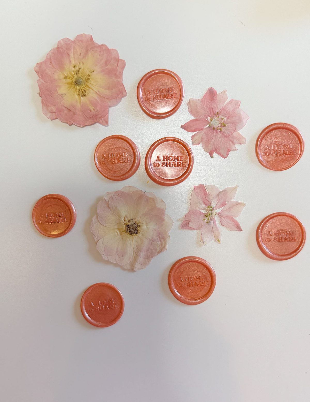 Pressed pink flowers and orange wax seals with the phrase "A HOME TO SHARE" embossed, arranged on a white background.