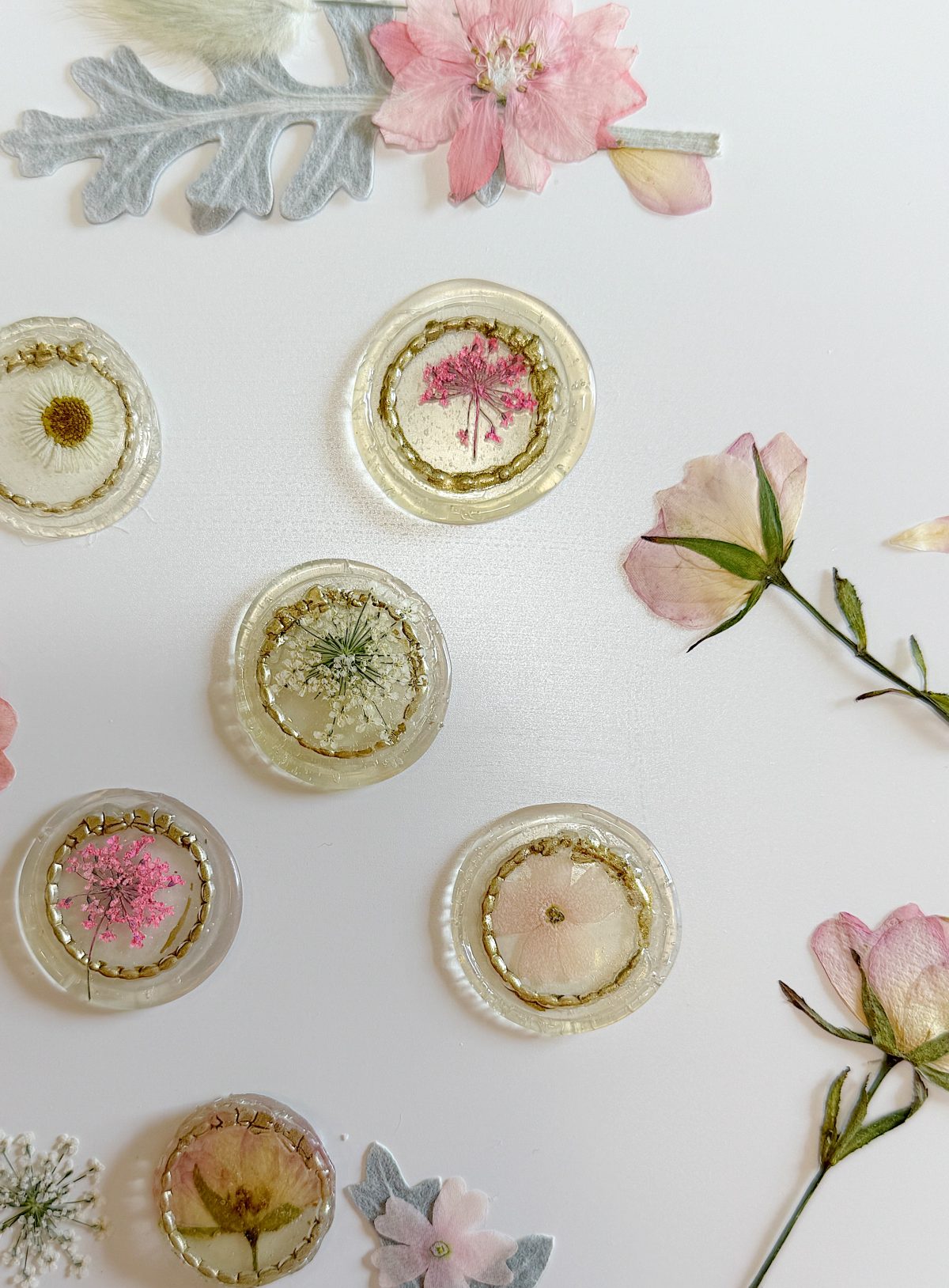 Clear resin circles with dried flowers inside are arranged on a white surface, surrounded by additional loose dried flowers and petals.