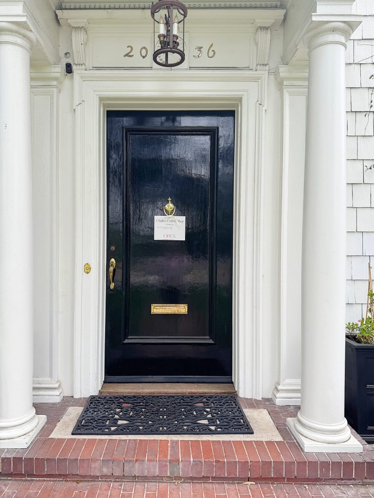 A black door with a brass knocker and mail slot, flanked by white columns. A sign on the door reads &ldquo;Please Come In. OPEN.&rdquo; The house number &ldquo;2056&rdquo; is above the door.