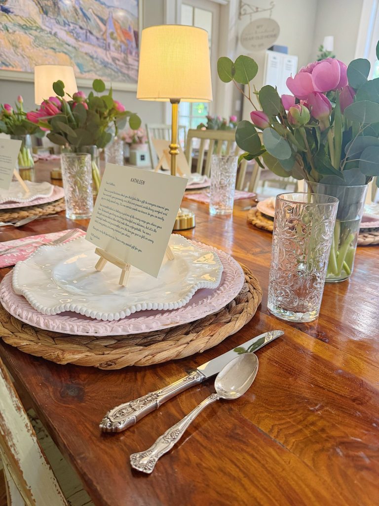 A wooden dining table set with plates, glasses, silverware, pink floral centerpieces, and small lamps, with printed menus displayed on mini easels at each place setting.