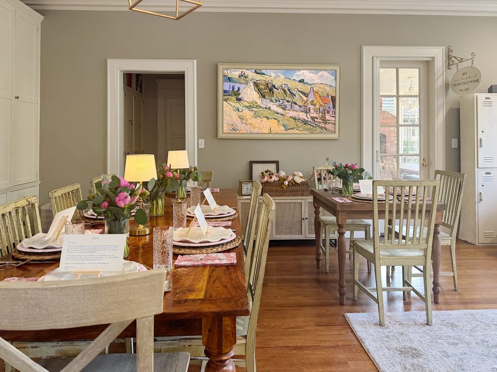 A dining room with two wooden tables set for a meal, decorated with flowers, place cards, and lamps, with a landscape painting on the wall and natural light from windows.