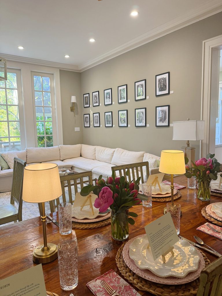 A dining table set with plates, glasses, flowers, and lamps is in the foreground; a white sectional sofa and framed black-and-white photos are in the background.