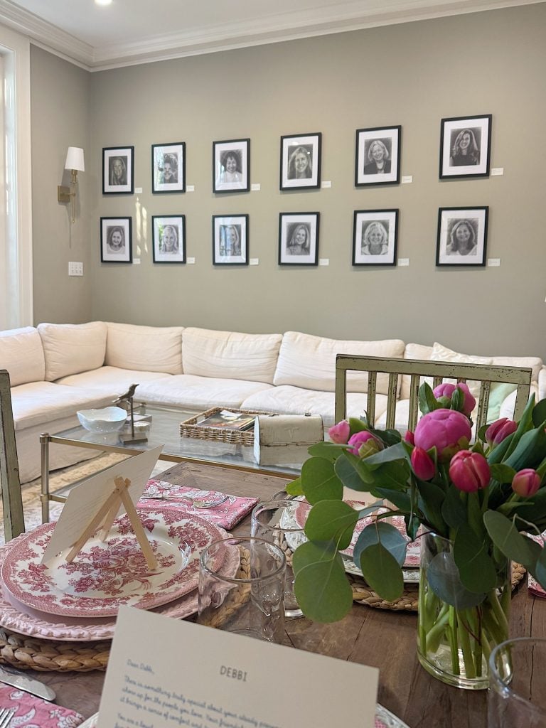 A living room with a white sectional sofa, framed black-and-white portraits on the wall, and a dining table set with pink tulips, pink plates, and a printed letter.