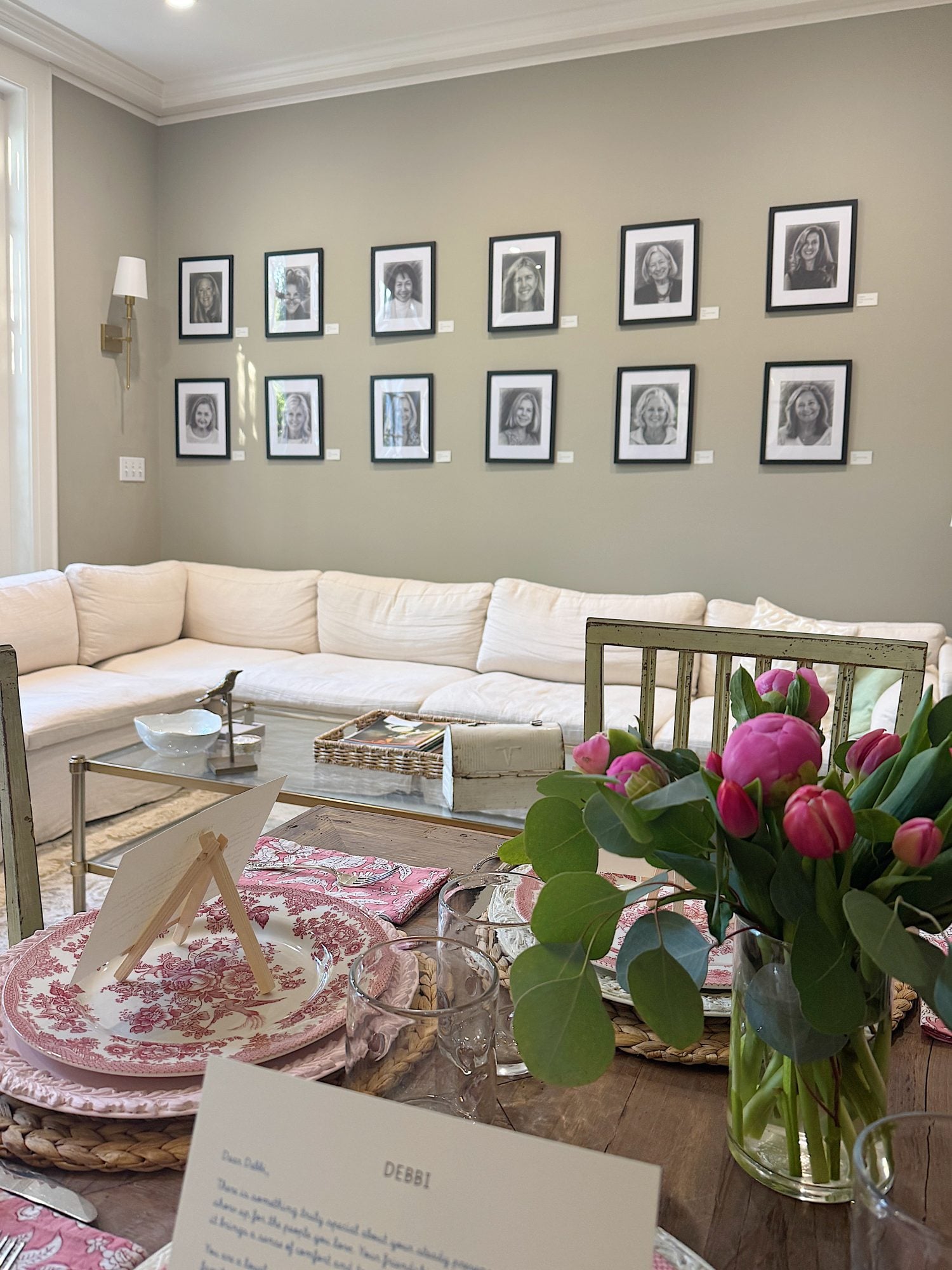 A living room with a white sectional sofa, framed black-and-white portraits on the wall, and a dining table set with pink tulips, pink plates, and a printed letter.