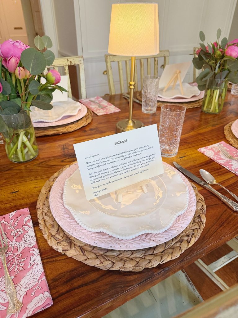 A wooden dining table is set with pink plates, woven placemats, pink floral napkins, glasses, and floral centerpieces. A lamp and a printed note sit at one place setting.