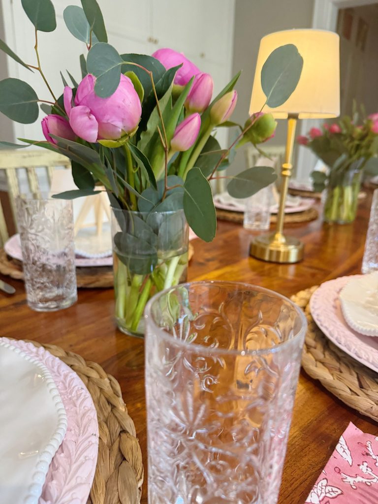 A wooden dining table set with glassware, white plates on woven placemats, a brass lamp, and a vase of pink flowers with green leaves.