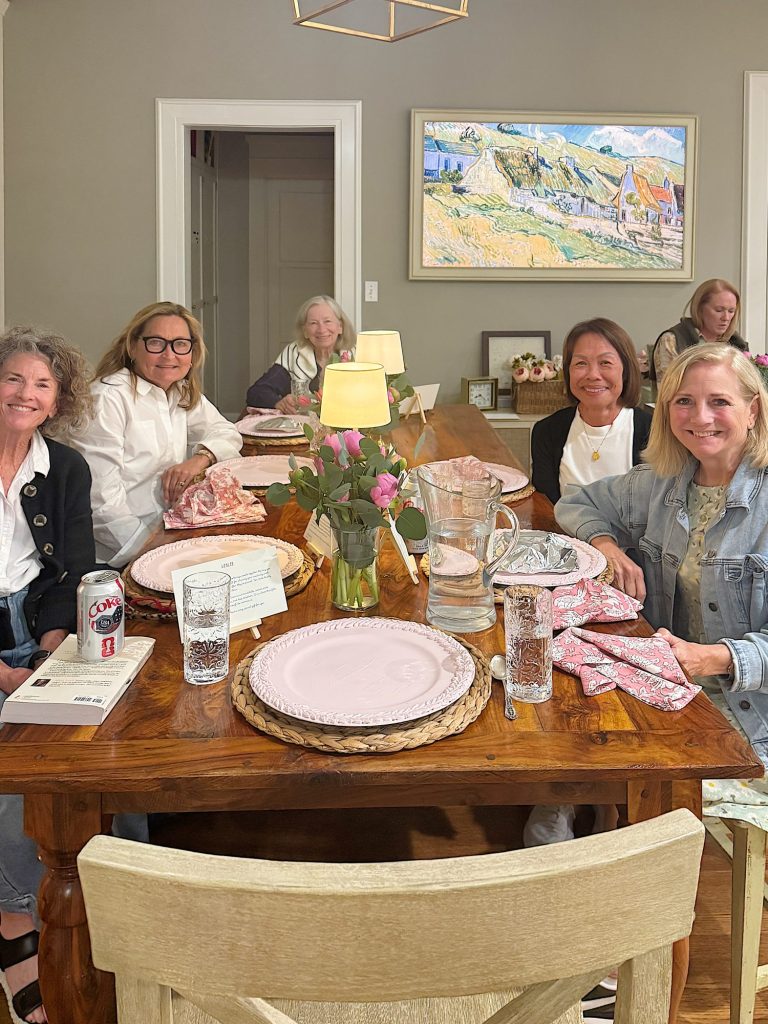 Six women sit around a dining table set with pink plates, glasses, and floral napkins. A vase with flowers and a pitcher of water are on the table; artwork hangs on the wall behind them.