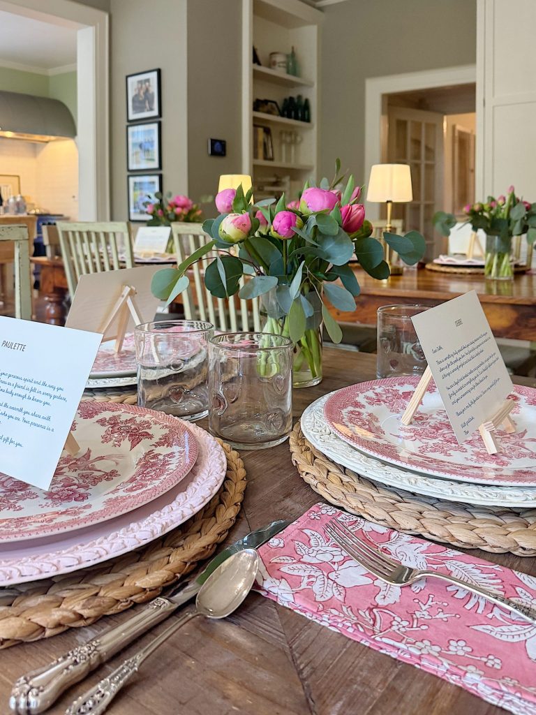 A wooden dining table is set with patterned plates, glassware, cutlery, pink floral napkins, menus on card holders, and a centerpiece of pink and green flowers.