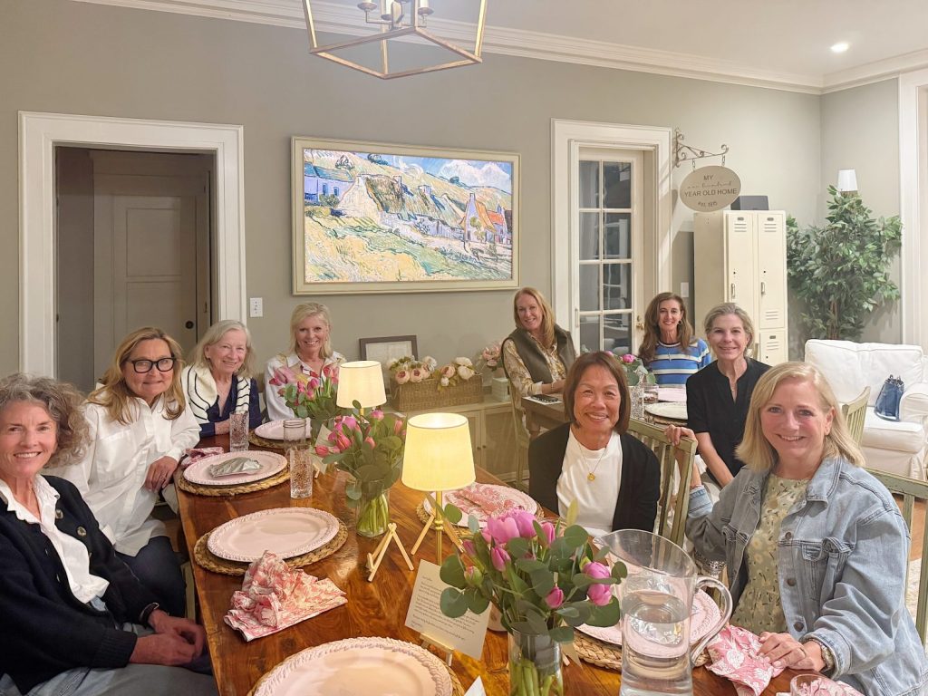 Nine women are seated around a wooden dining table set with pink plates, napkins, flowers, and lamps in a well-lit living room.