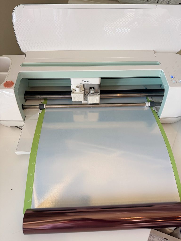 A Cricut cutting machine in use, with a metallic silver sheet loaded on a green cutting mat, ready for a craft or design project.