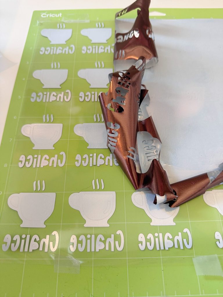 A green Cricut cutting mat holds white vinyl cutouts of cups and the word &ldquo;Chai&rdquo; with a brown sheet of excess vinyl partially removed and curled up.