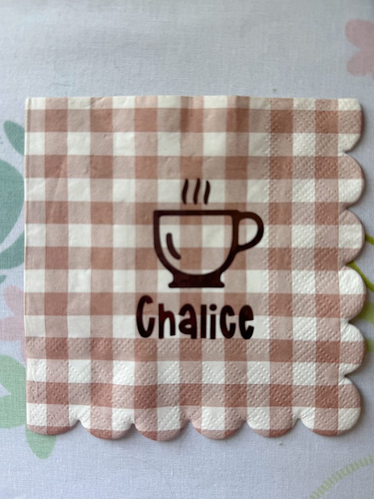 A brown and white checkered napkin with a scalloped edge, featuring an illustration of a steaming mug and the word "Chalice" printed below it.