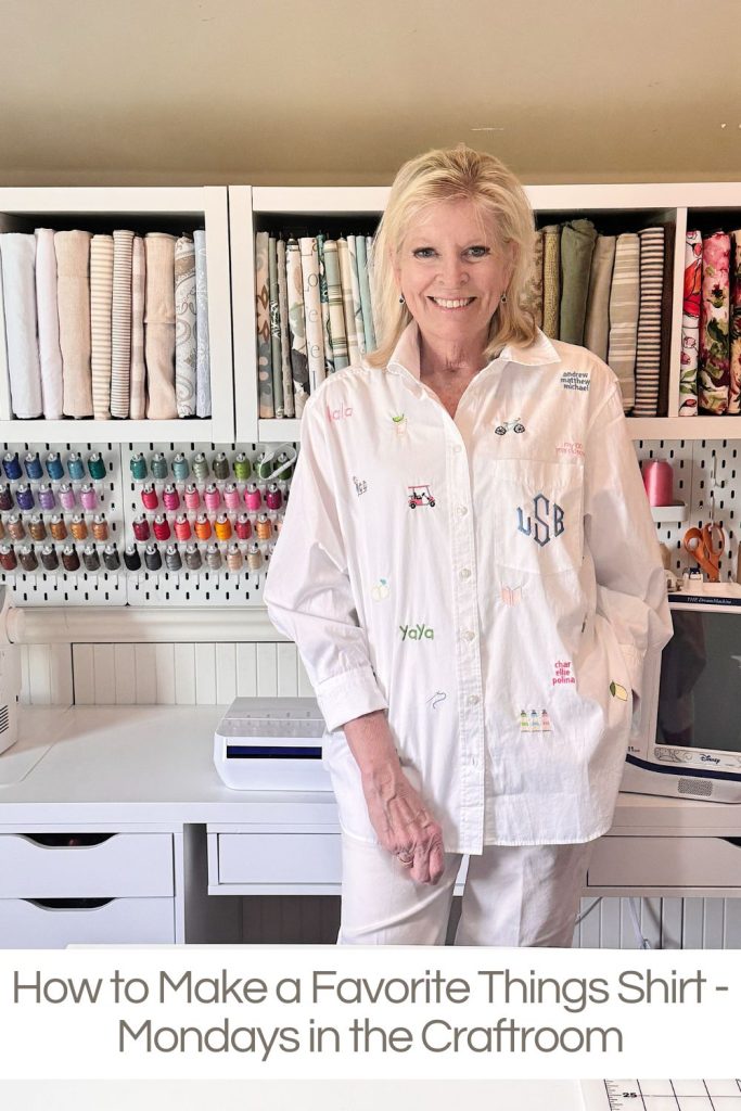 A woman stands in a craft room wearing a white shirt with various colorful embroidered designs, surrounded by shelves of fabric and thread.