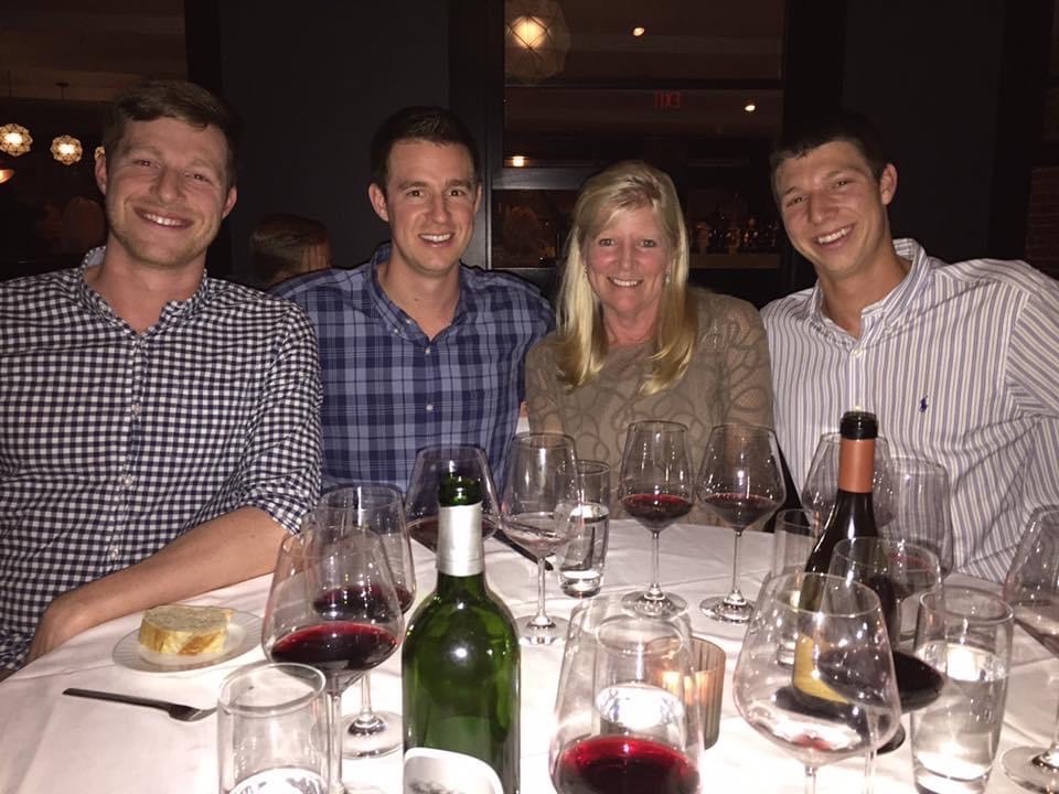 Four people sit at a restaurant table with multiple glasses of red wine, a wine bottle, and a bread plate in front of them, smiling at the camera.