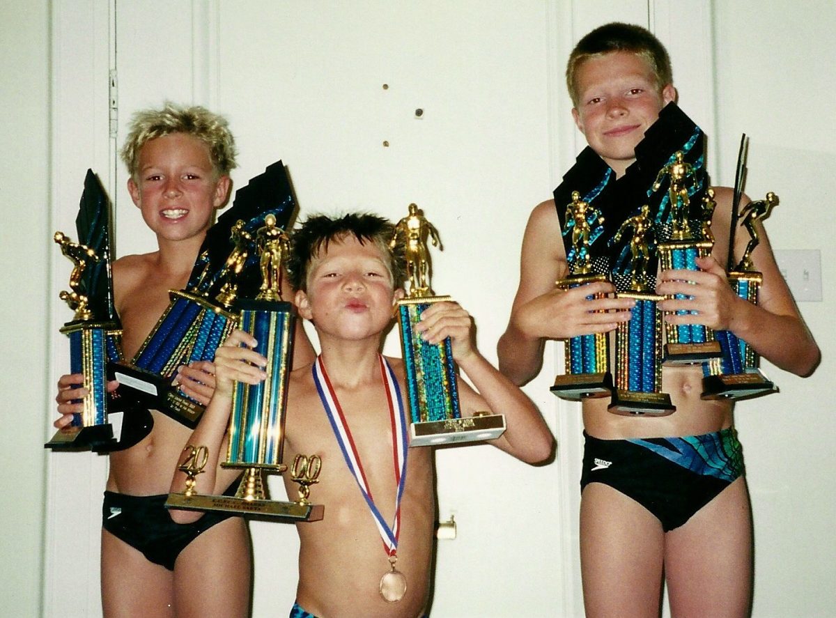 Three boys in swim trunks hold multiple trophies and one wears a medal, standing in front of a white door and smiling at the camera.