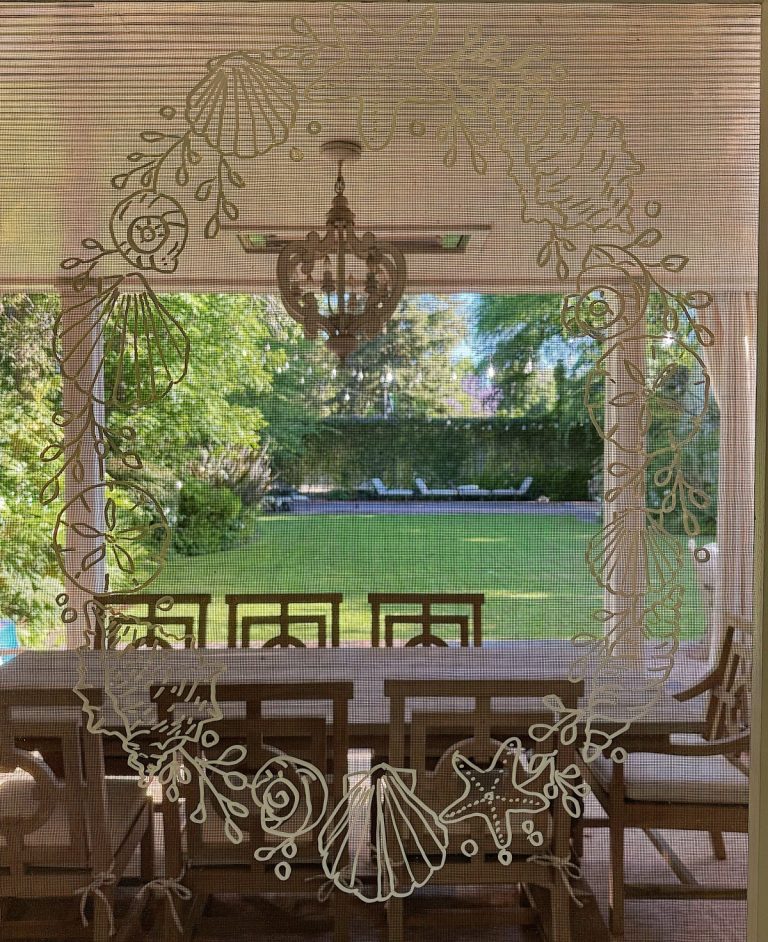 A dining table set with chairs is seen through a window decorated with white sea shell and starfish drawings, overlooking a green backyard with trees and lounge chairs.