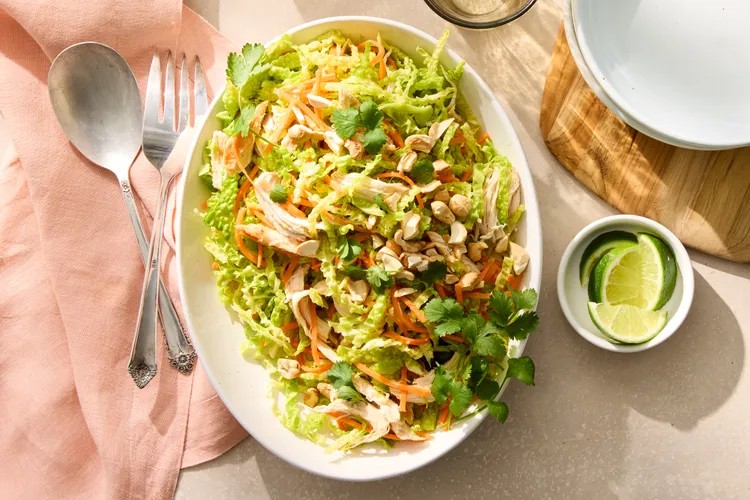 A white oval bowl filled with shredded chicken salad with lettuce, carrots, cilantro, and chopped nuts, with lime wedges and a set of silverware nearby.