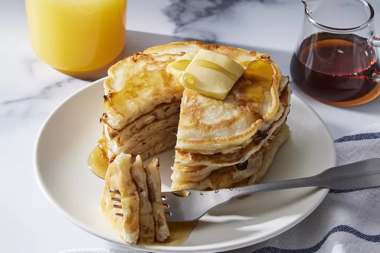 A stack of pancakes topped with butter and syrup sits on a white plate, with a fork holding a bite. A glass of orange juice and a jar of syrup are nearby.