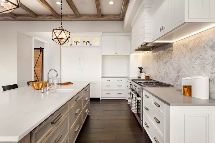 Modern kitchen with white cabinets, marble backsplash, stainless steel appliances, and a large island with pendant lighting and wooden accents on the ceiling.