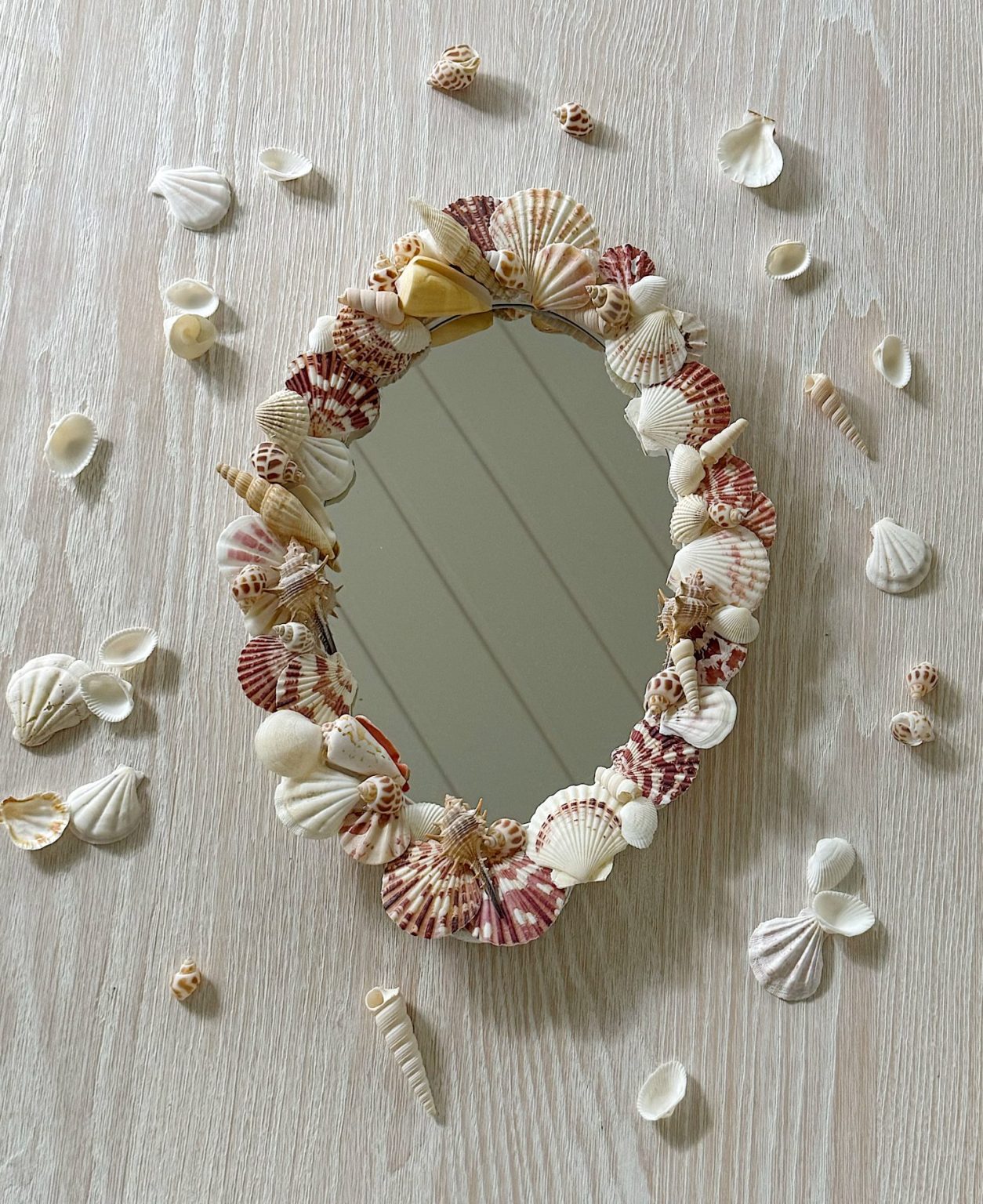 Oval mirror decorated with assorted seashells, placed on a light wooden surface with additional seashells scattered around it.