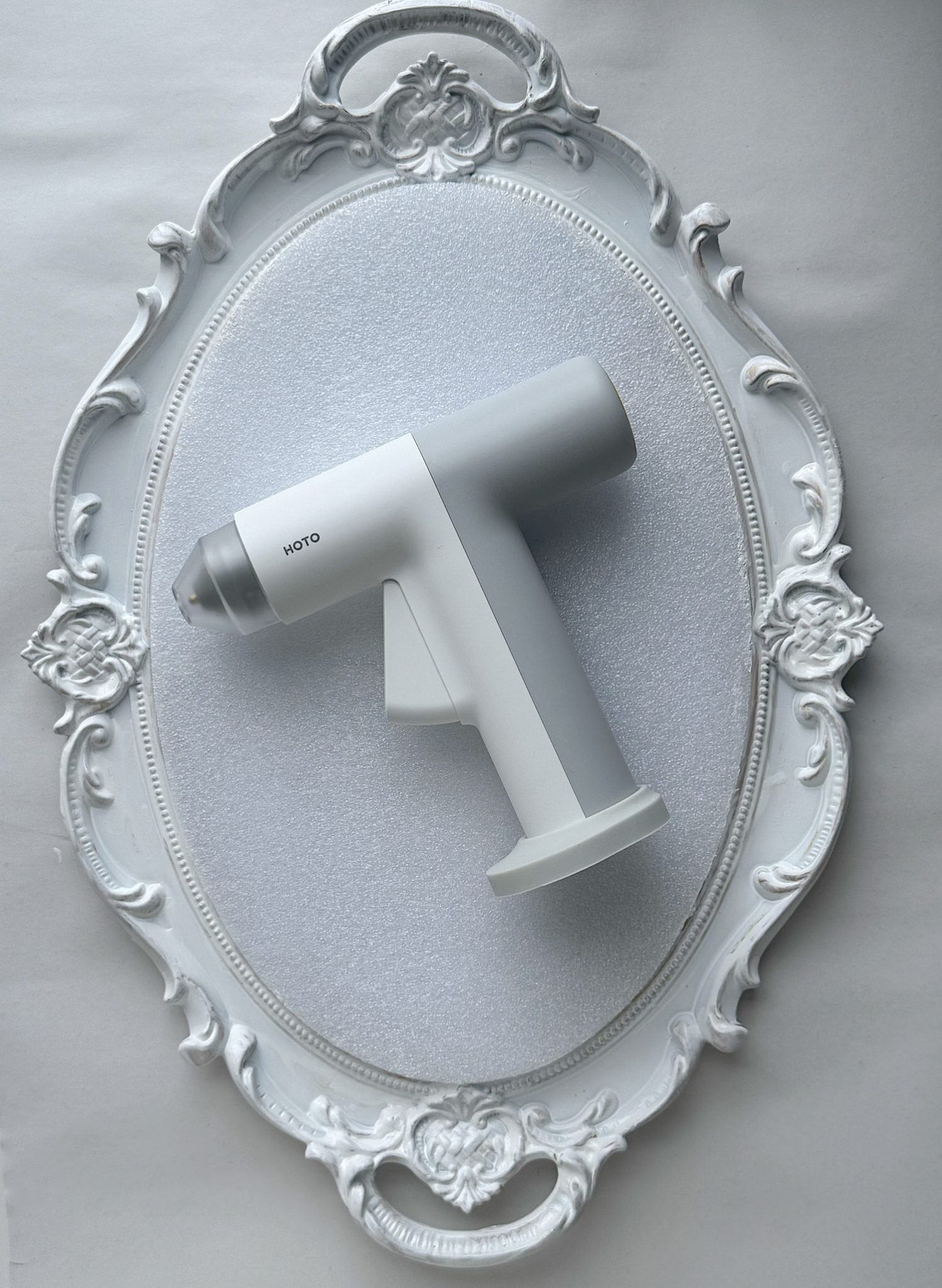 A white handheld device labeled "KOTO" is placed on an ornate white oval mirror with decorative floral details.