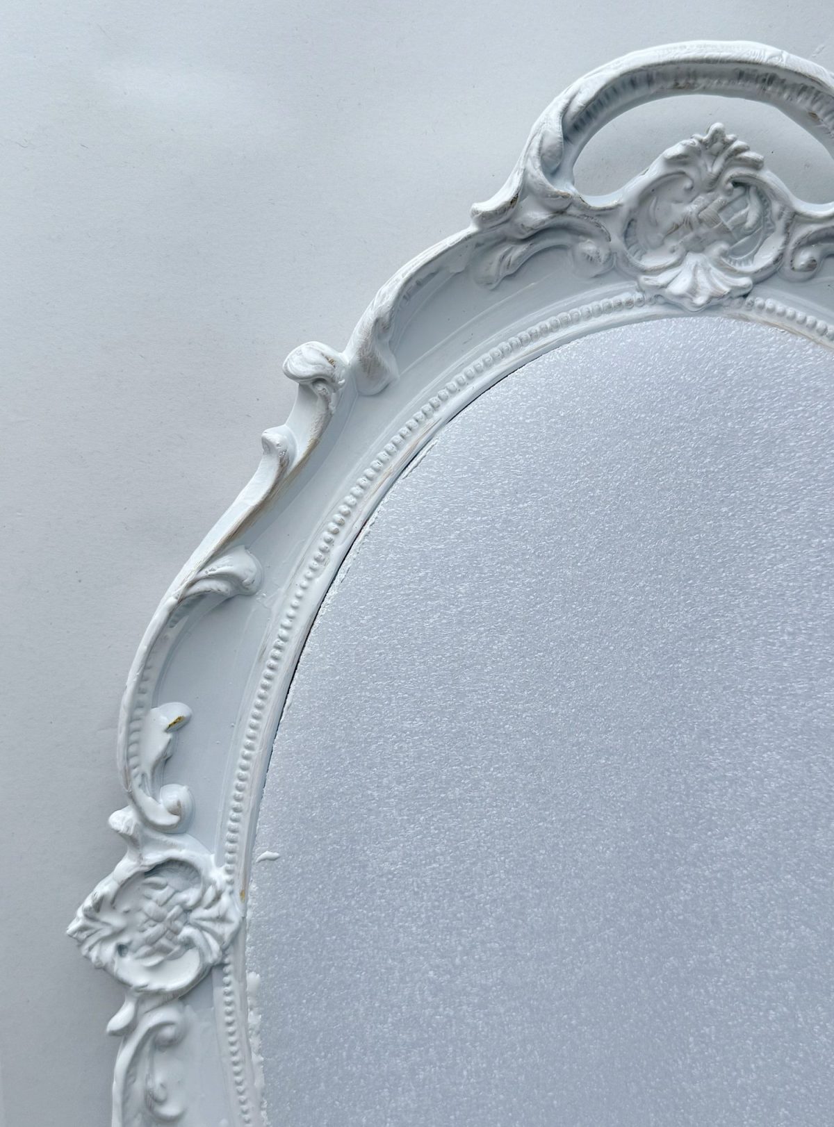Close-up of the ornate top left corner of a white decorative oval mirror frame against a plain white background.